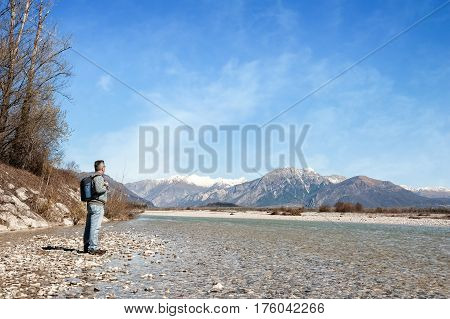 Mature Hiker On The Bank Of A River. Walking Toward Mountain.