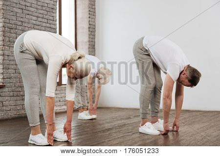 Stretch your back. Three friends wearing white sneakers standing in semi position while touching wooden floor