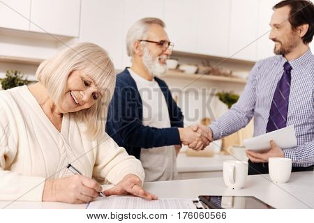 The moment of signing important documents. Charming delightful smiling aged woman sitting and signing contract while her husband greeting the lawyer in the background
