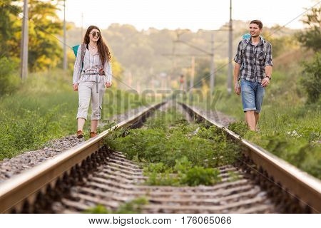 Two jolly Hikers Man and Girl in casual Travel Clothing with Backpacks walking along old Country Railroad with back light Sun and Forest on Background