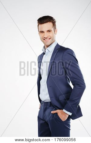 Vertical image of smiling business man which standing sideways with arms in pockets and looking at camera. Isolated white background