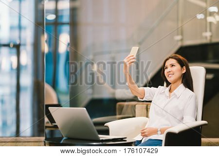 Beautiful Asian woman selfie or VDO call on smartphone working with laptop in office or living room. Modern lifestyle with gadget technology or casual business concept