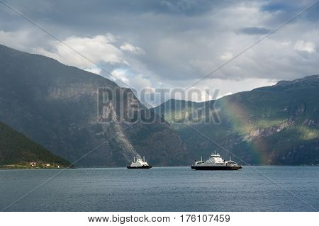 Landscape photo of norwegian fjord with two ferries. Cloudy weather after the rain, rare sun beams and rainbow. Sognefjorden, Norway.