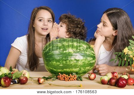 Small curly boy is telling a secret girls. Sitting at the table in front of a large watermelon and fruit. Blue background. Close-up.