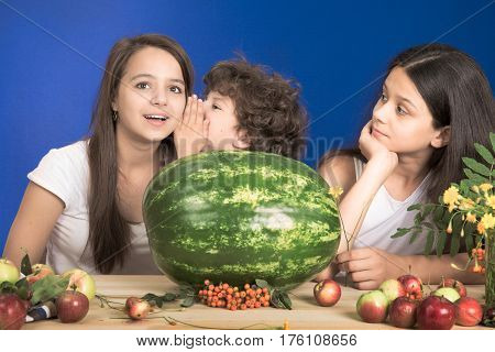 Children tell secrets to each other sitting at the table. Two girls and a boy. Blue background. Close-up.