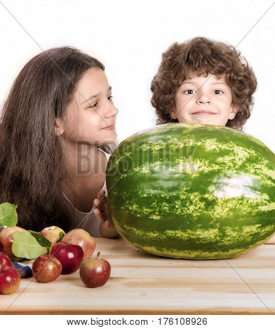 Little curly boy and a girl sitting at a wooden table and look at the big whole watermelon. White background. Close-up.