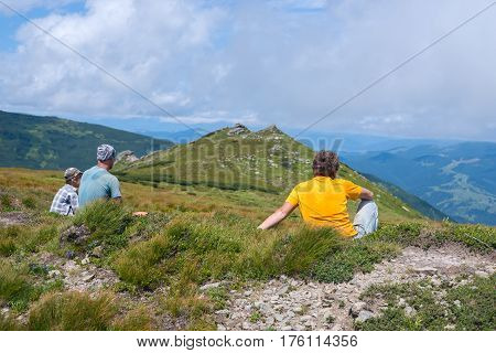Hiker man sitting on an alpine meadow next to his friends. Back view. Sunny day in the mountains.