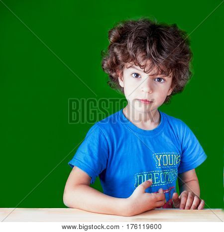 Cute curly little boy put his hand on the table and looking thoughtfully into the camera. Sitting at a wooden table. On a green background.