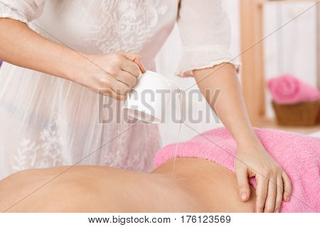 Closeup of masseur hands holding  massage heating oil pouring oil on woman's back. Beauty care