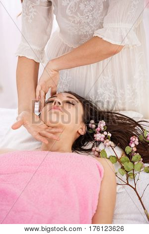 Masseur hands doing facial massage carefully on young lady lying on couch with tender flowers
