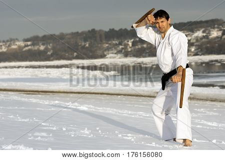 The man in white kimono is practicing japanese martial arts with tonfa in the snow on winter background.