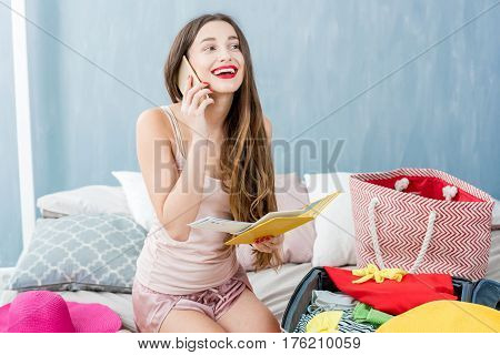 Young woman calling with phone sitting with suitcase and clothes on the bed. Prepairing for the summer vacations