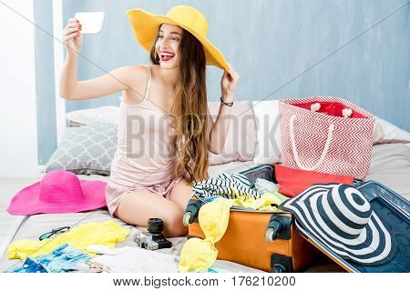 Young woman choosing hat sitting with clothes and suitcase on the bed. Prepairing for the summer vacations