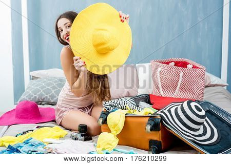 Young woman choosing hat sitting with clothes and suitcase on the bed. Prepairing for the summer vacations