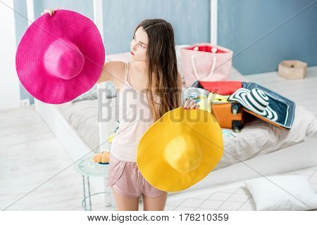 Young woman choosing hat in the bedroom with suitcase and clothes on the background. Prepairing for the summer vacations