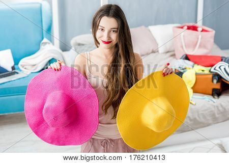 Young woman choosing hat in the bedroom with suitcase and clothes on the background. Prepairing for the summer vacations