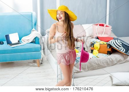 Young woman choosing hat in the bedroom with suitcase and clothes on the background. Prepairing for the summer vacations