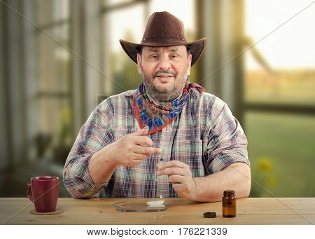 Middle-aged farmer in brown hat makes sure that all air is out of the syringe before delivering medication for himself. Bearded cowboy sits at wooden desk looking directly at camera. There are maroon mug, small bottle with rubbing alcohol on the table. Ho