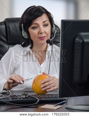 Telemedicine female doctor in headset guides an intramuscular injection by internet.  Mature woman sits opposite desktop monitor and makes clear to someone technique with syringe and grapefruit. Vertical mid-shot on blurred indoors background