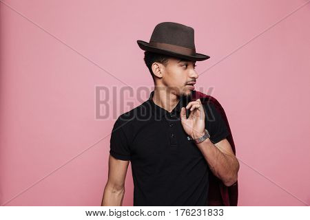 Portrait of a young handsome afro american man in hat holding jacket over shoulder isolated on the pink background
