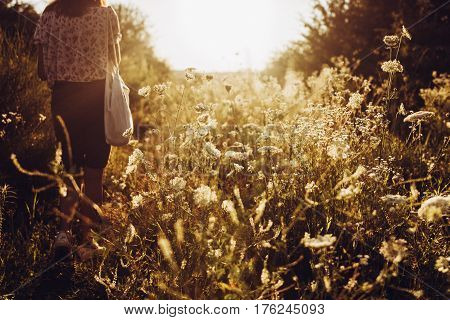 Stylish Hipster Woman With Eco Bag Walking Into Sunlight Among Wildflowers In Summer Evening Field U