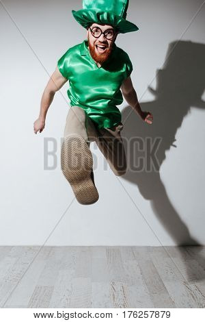 Vertical image of happy man in st.patriks costume and eyeglasses which jumping in studio. Isolated gray background