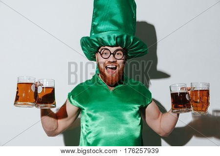 Happy guy in st.patriks costume and eyeglasses which holding many cups of beer and looking at camera. Isolated gray background