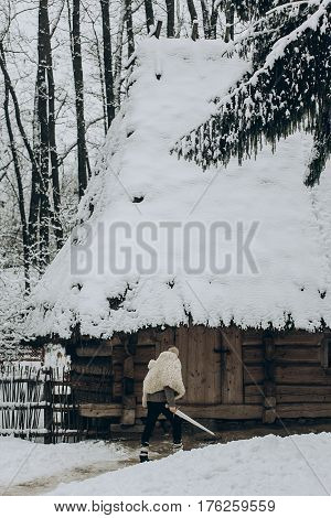 Strong Viking With Sword And Wolf Pelt Walking Towards Historical Wooden Building In North Woods In