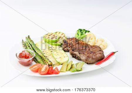 Grilled beef grilled with asparagus, zucchini, lobi, broccoli, tomato, chili and red sauce on a white plate, over white background