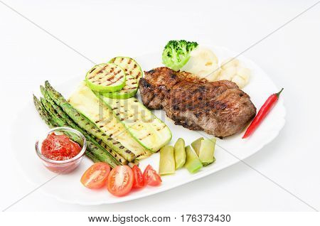 Grilled beef grilled with asparagus, zucchini, lobi, broccoli, tomato, chili and red sauce on a white plate, over white background