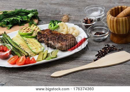 Grilled beef grilled with asparagus, zucchini, lobi, broccoli, tomato, chili and red sauce on a white plate, and other vegetables in wood background