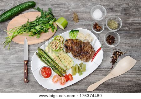 Grilled beef grilled with asparagus, zucchini, lobi, broccoli, tomato, chili and red sauce on a white plate, and other vegetables in wood background