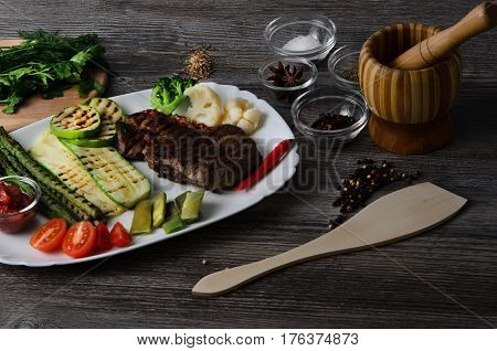 Grilled beef grilled with asparagus, zucchini, lobi, broccoli, tomato, chili and red sauce on a white plate, and other vegetables in wood background