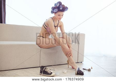 Beautiful girl in hair curlers in sexy lacy lingerie putting on fishnet stockings. Attractive female dressing up on white. Undressed woman sitting on sofa at home and preparing on party. Make up accessories on the floor