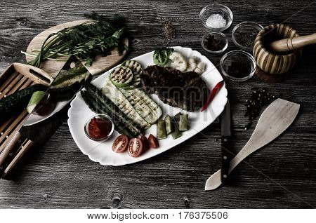 Grilled beef grilled with asparagus, zucchini, lobi, broccoli, tomato, chili and red sauce on a white plate, and other vegetables in wood background