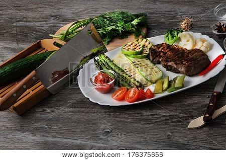 Grilled beef grilled with asparagus, zucchini, lobi, broccoli, tomato, chili and red sauce on a white plate, and other vegetables in wood background