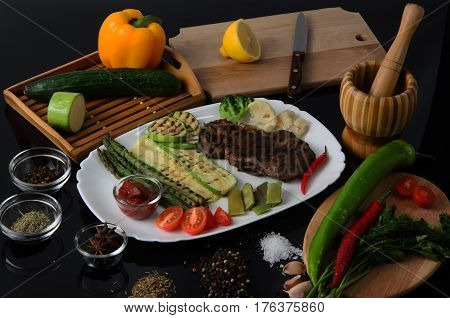 Grilled beef grilled steak with asparagus, zucchini, lobi, broccoli, tomato, chili and red sauce on a white plate, on a black glossy background