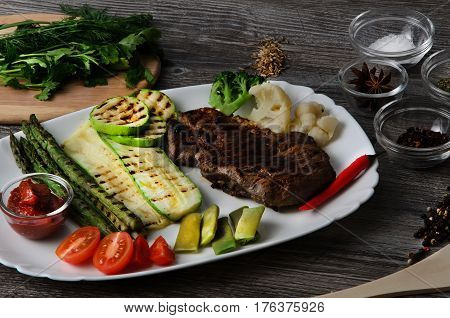 Grilled beef grilled with asparagus, zucchini, lobi, broccoli, tomato, chili and red sauce on a white plate, and other vegetables in wood background