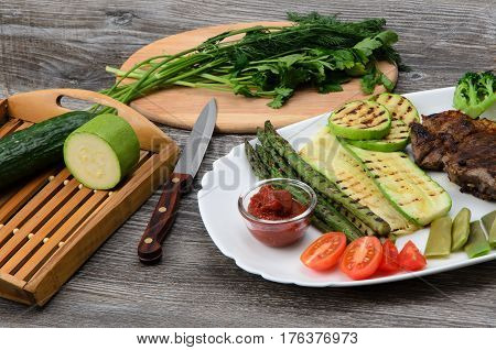 Grilled beef grilled with asparagus, zucchini, lobi, broccoli, tomato, chili and red sauce on a white plate, and other vegetables in wood background