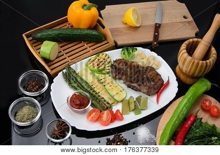 Grilled beef grilled steak with asparagus, zucchini, lobi, broccoli, tomato, chili and red sauce on a white plate, on a black glossy background