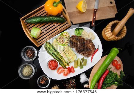 Grilled beef grilled steak with asparagus, zucchini, lobi, broccoli, tomato, chili and red sauce on a white plate, on a black glossy background
