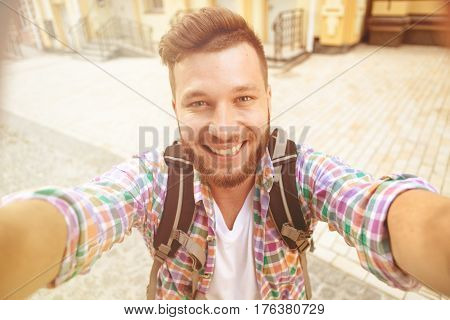 Smiling tourist with backpack making selfies isolated on beautiful modern buildings located in the city centre. Man happy spending his daytime. Toned image.