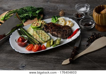 Grilled beef grilled with asparagus, zucchini, lobi, broccoli, tomato, chili and red sauce on a white plate, and other vegetables in wood background