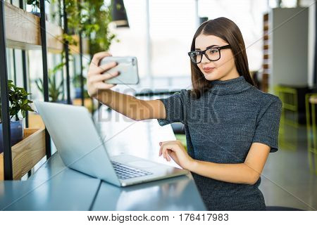 Young female taking a selfie in the office while sitting with laptop in modern office room