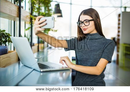 Young female taking a selfie in the office while sitting with laptop in modern office room