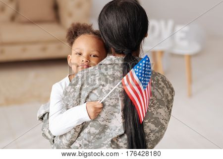 Glad to have you here. Lovely charming pretty girl hugging her mom tightly while greeting her home and holding a flag in her hand