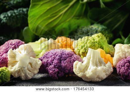 Variety of raw green vegetables salads, lettuce, bok choy, corn, broccoli, savoy cabbage, colorful young cauliflower over black stone texture background. Close up