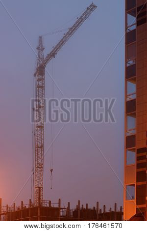 Construction crane on the site, unfinished house, fog covers the upper floors, evening twilight.