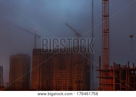 Two cranes on the construction site, unfinished house, foggy evening twilight.