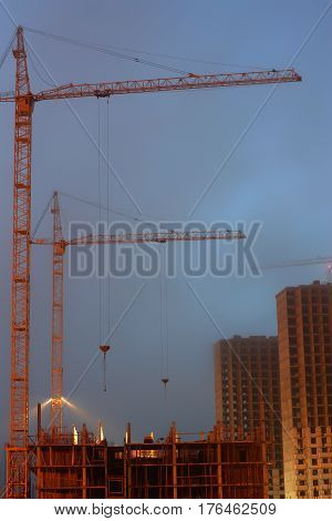 Two cranes on the construction site, unfinished multi-storey house, foggy evening twilight, building lighting.